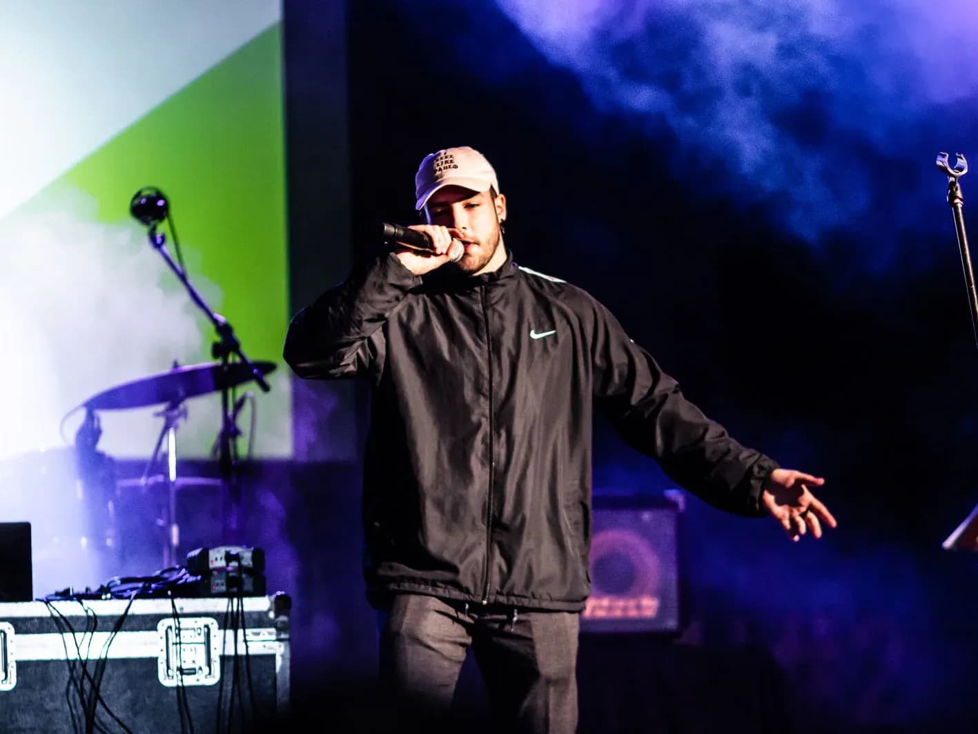 AkaEdu cantando con brazo extendido sobre escenario con iluminación verde y morada, actuación de música urbana en festival