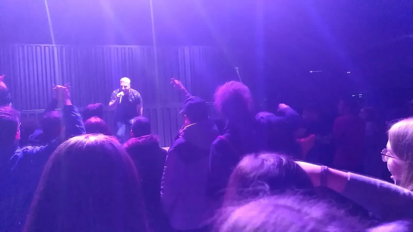AKA Edu rapeando con microfono sobre escenario bañado en luz violeta con publico levantando brazos en concierto nocturno