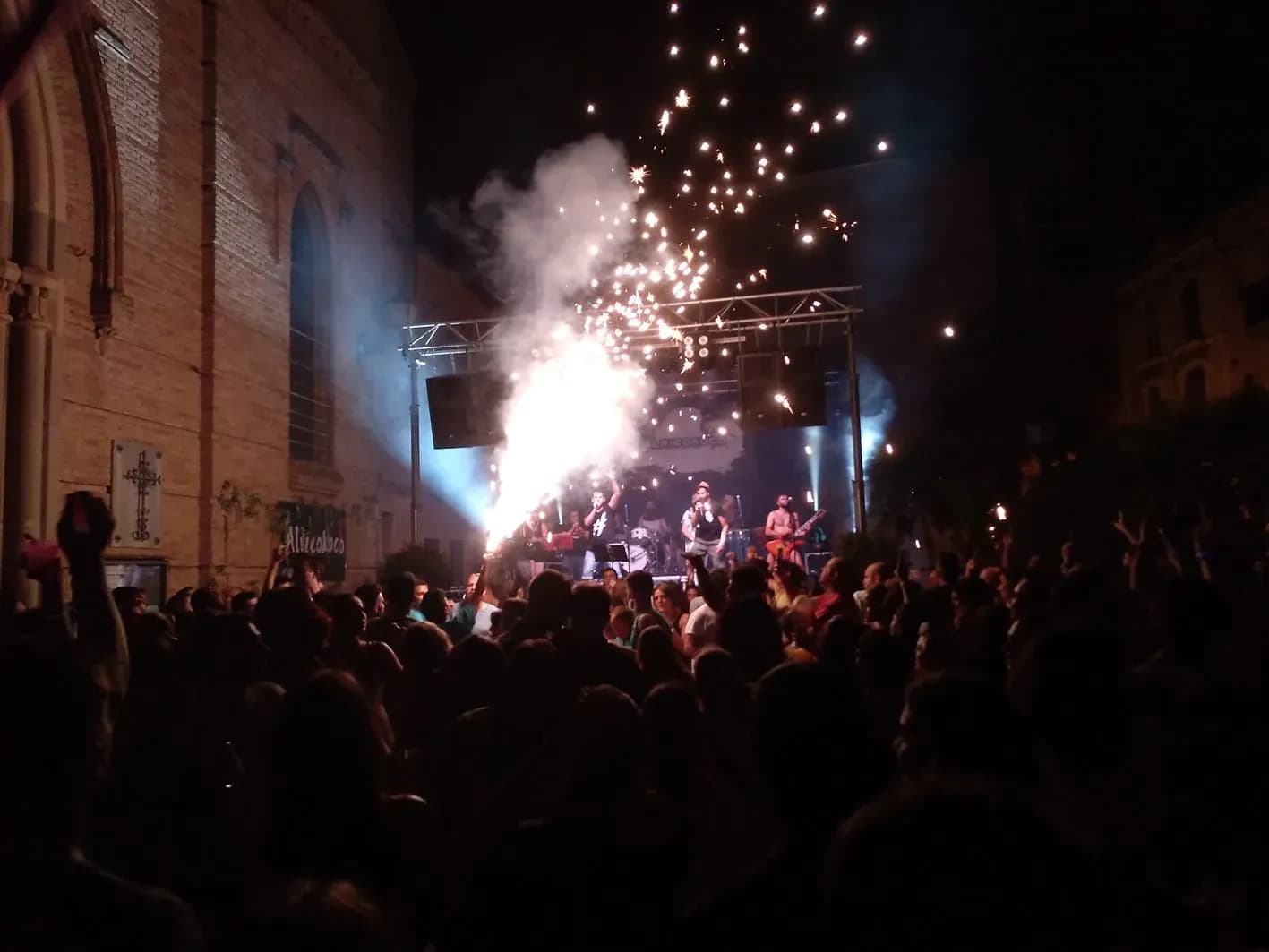Concierto de Alricokoco al aire libre con fuegos artificiales sobre el escenario y público levantando los brazos de noche