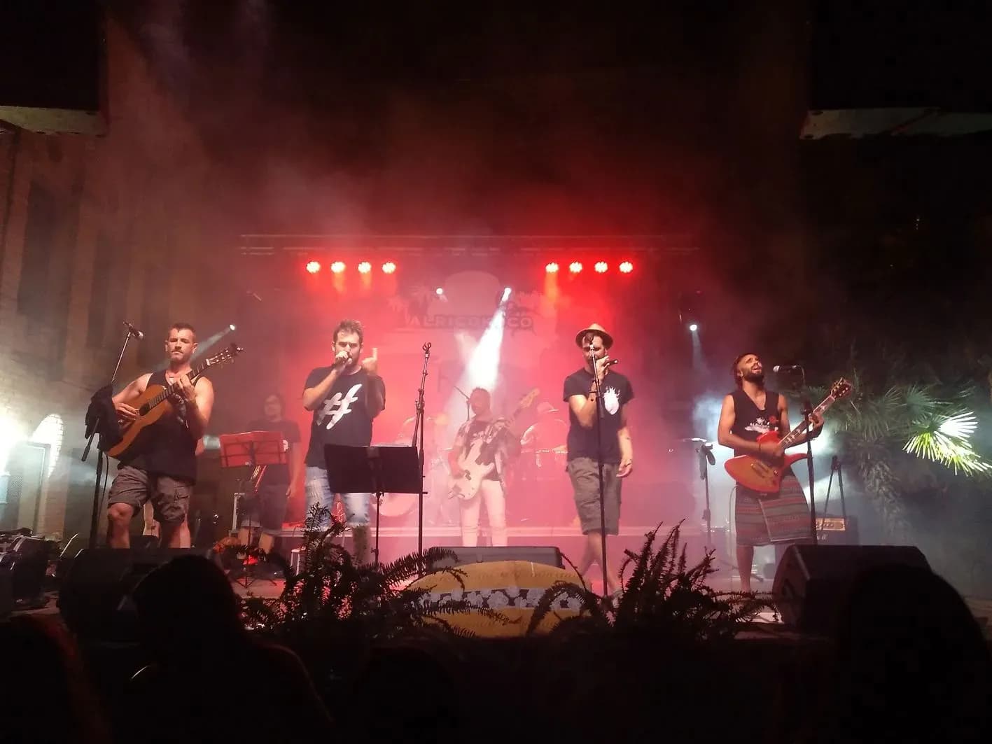 Alricokoco en directo con iluminación roja, cantantes y guitarras acústicas sobre escenario en fiesta mayor de pueblo