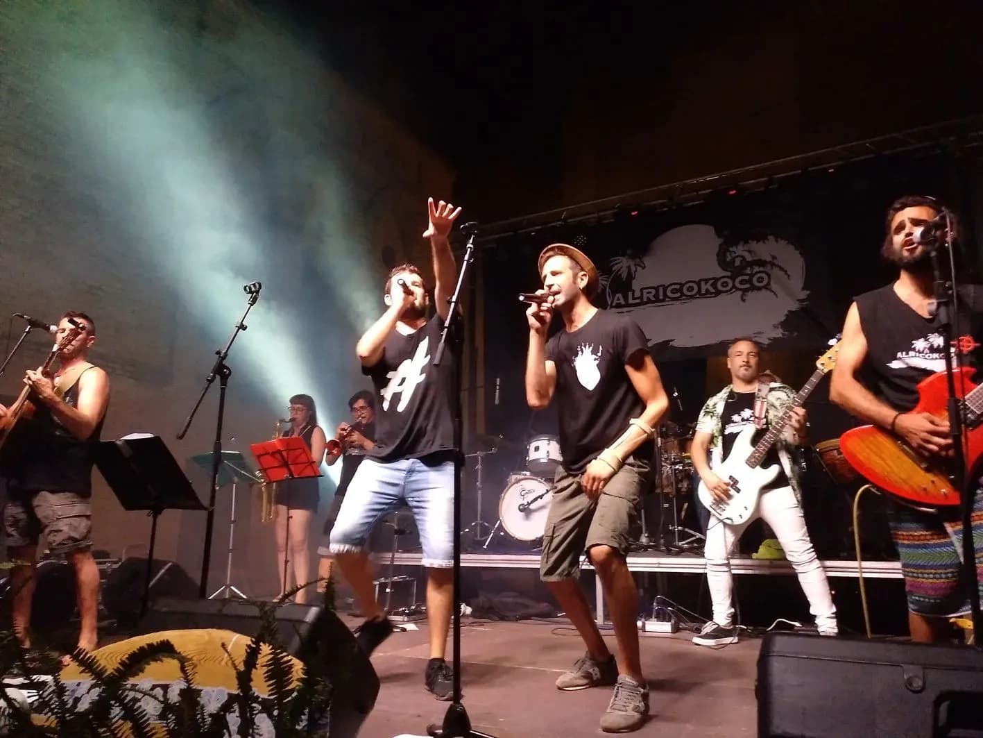 Vocalistas de Alricokoco animando al público con micrófonos y la banda al completo de fondo durante concierto en plaza