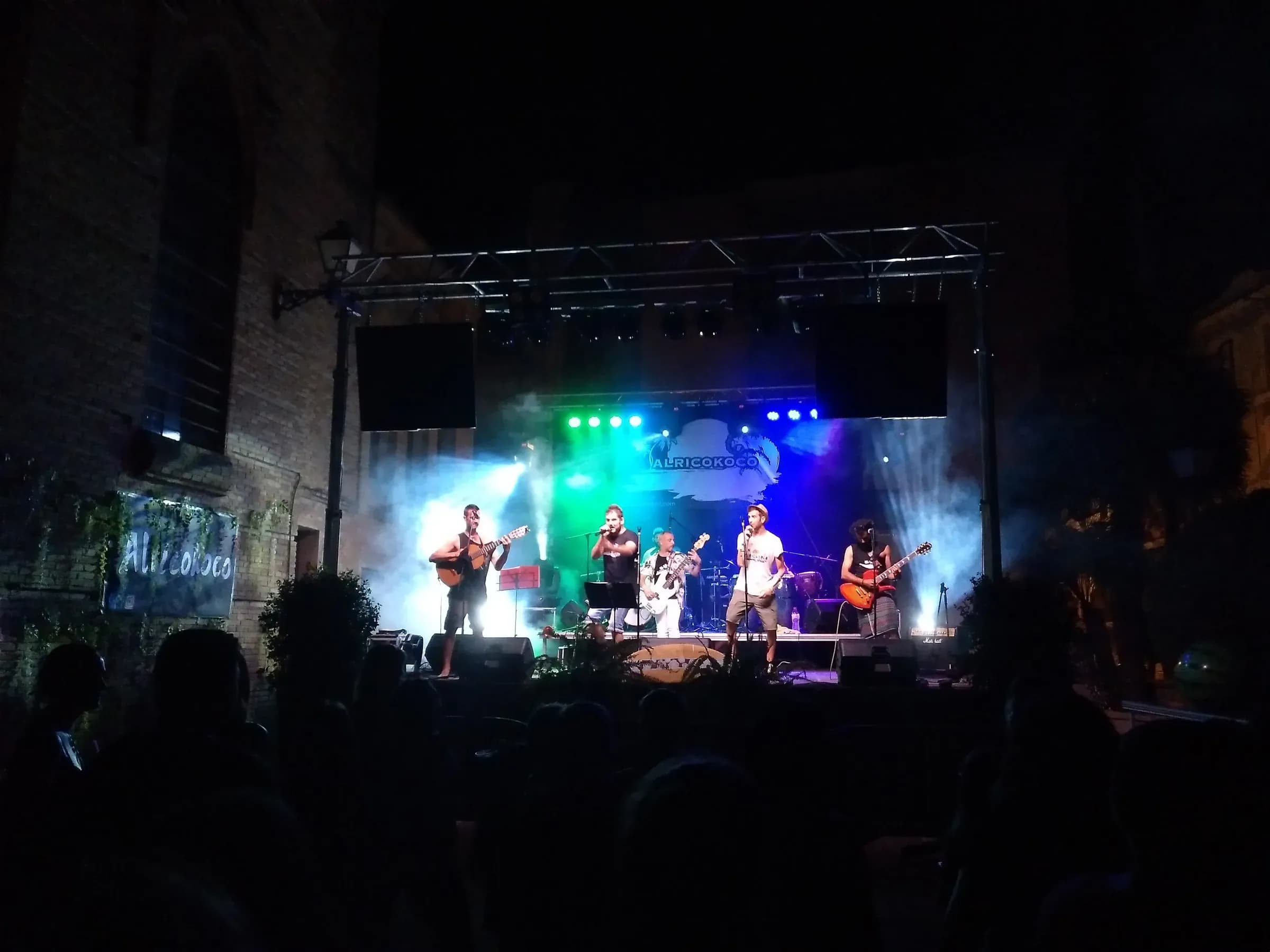 Banda Alricokoco en escenario nocturno con humo y luces verdes y azules ante publico en plaza con muros de piedra