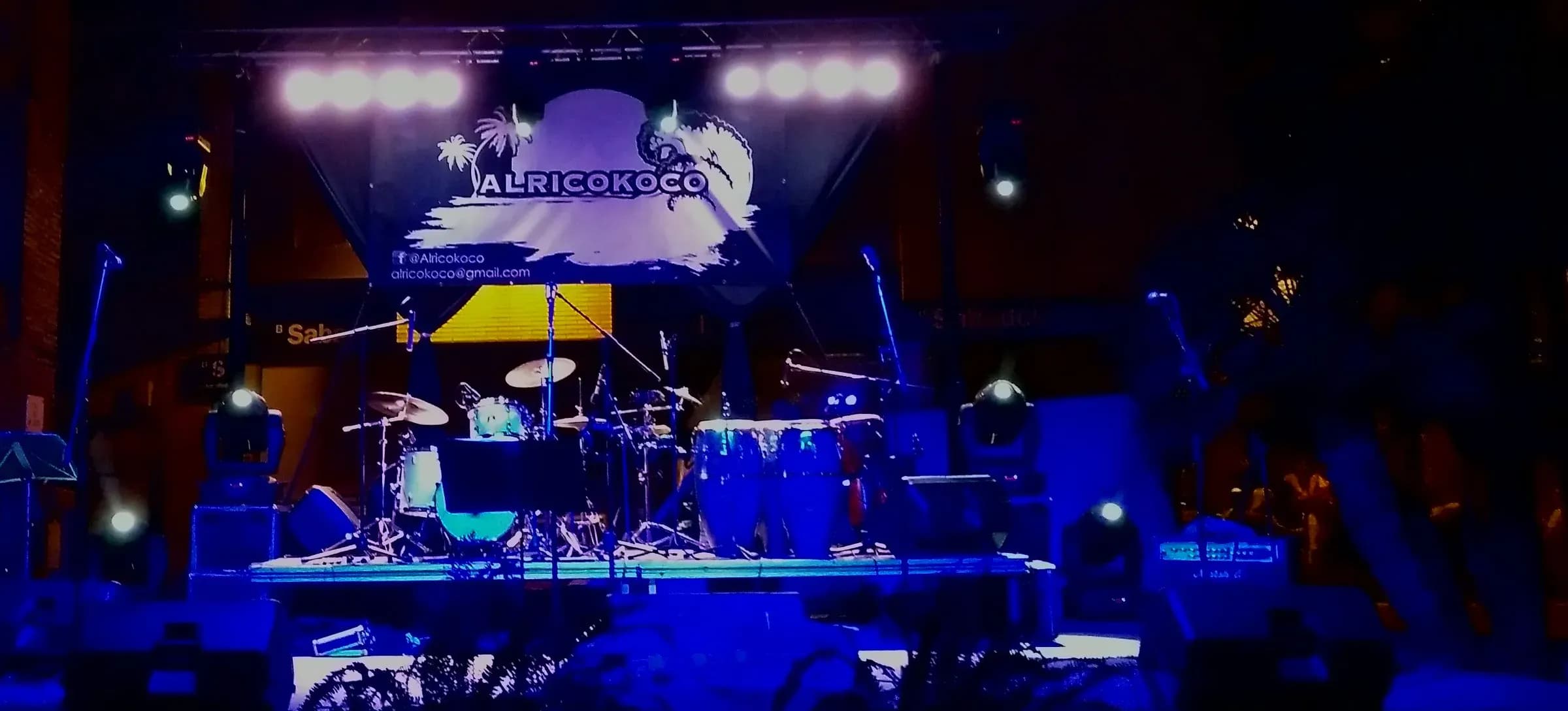 Escenario nocturno de Alricokoco con banner del grupo iluminado en azul y bateria preparada antes del concierto