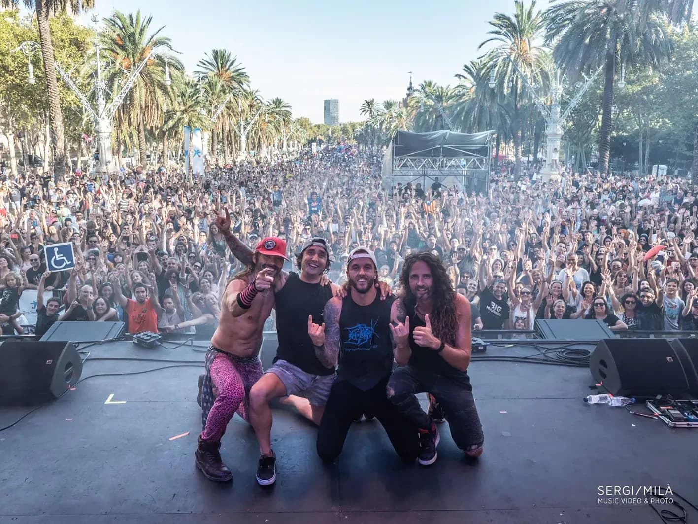 Big Mouthers al completo posando arrodillados sobre escenario con multitud y palmeras de fondo en concierto de Barcelona