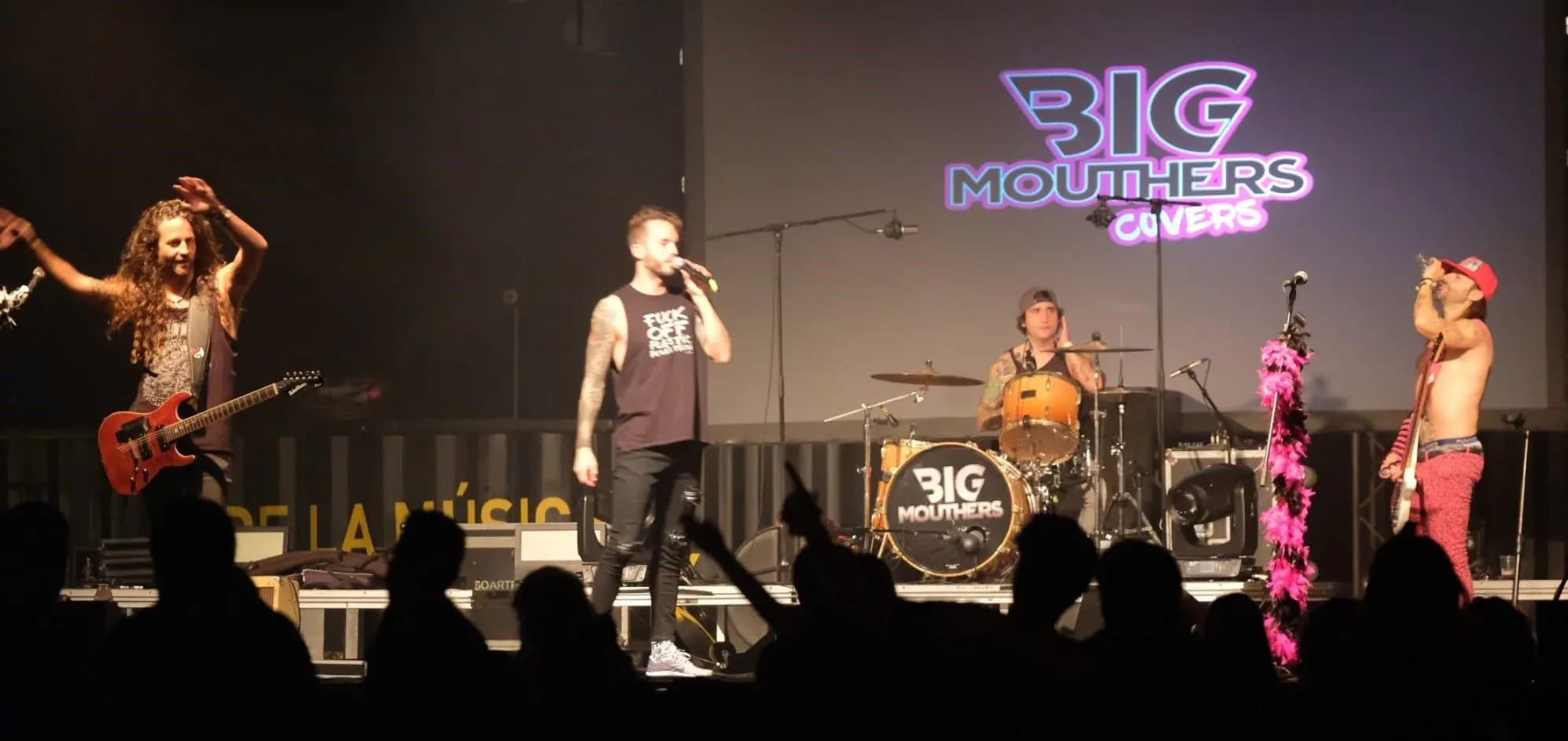 Big Mouthers en directo con cantante guitarrista bateria y publico en penumbra ante la pantalla con su logo
