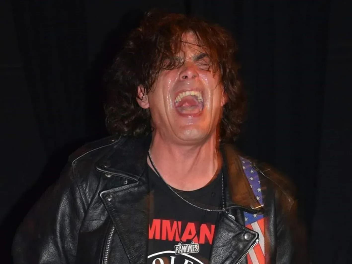Bajista de Commando con peluca rubia, chupa de cuero y bajo blanco junto a roadie con cartel Gabba Gabba Hey y batería logo