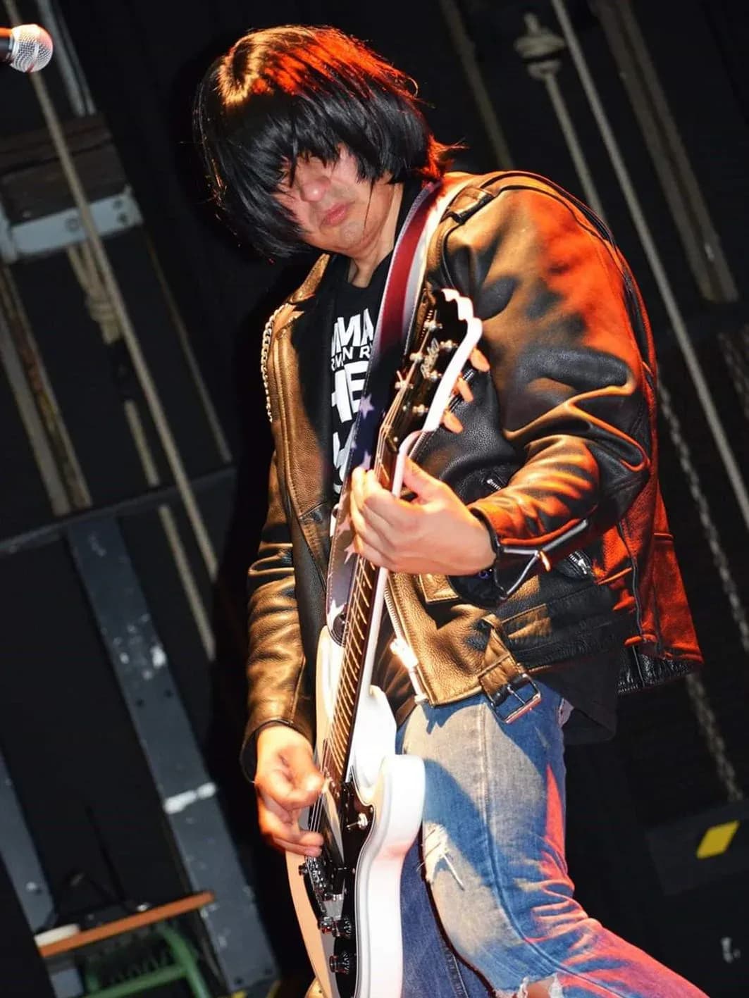 Guitarrista de Commando con flequillo cubriendo cara, chaqueta de cuero marron y guitarra blanca tocando en directo