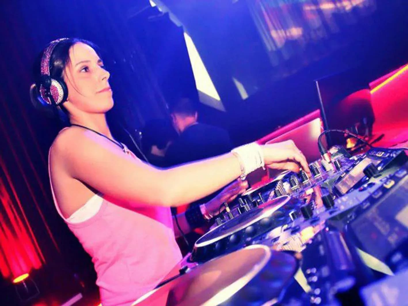 DJ Enka con auriculares plateados y top rosa manejando mesa de mezclas bajo luz roja de sala nocturna en Calafell