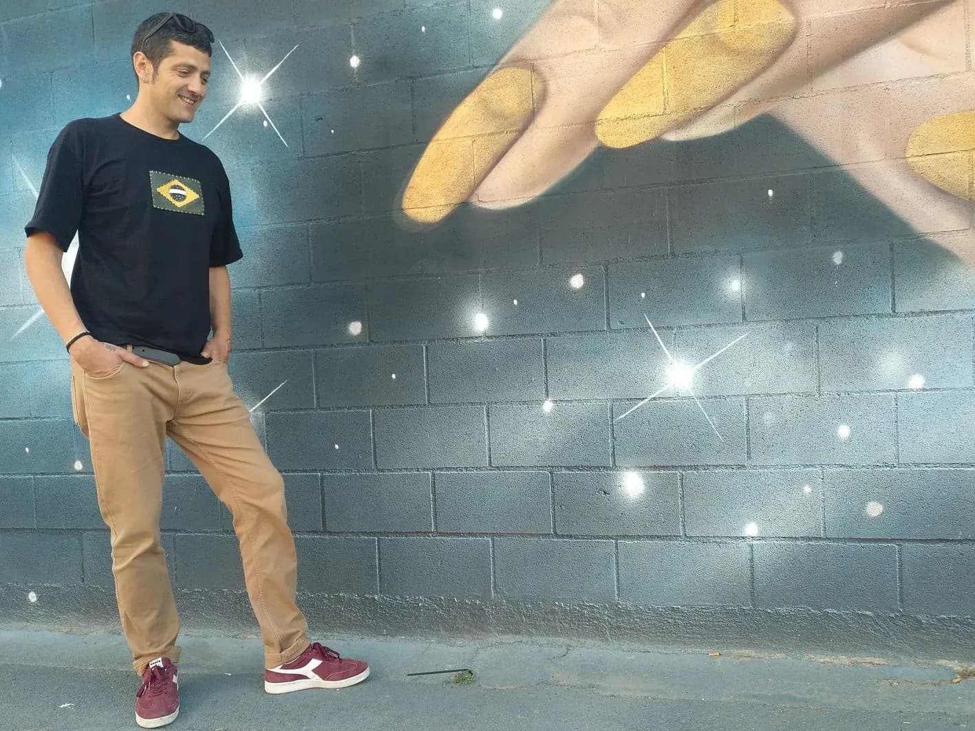 DJ Pablo de pie junto a mural de mano gigante con destellos brillantes posando en exterior con camiseta negra