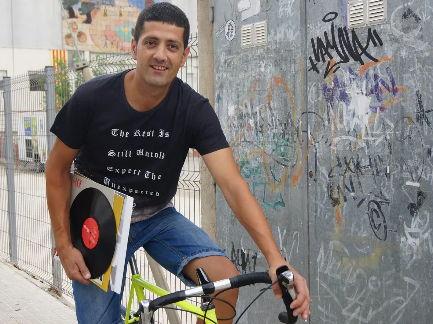 DJ Pablo apoyado en bicicleta sosteniendo vinilo, con camiseta negra y muro de grafitis de fondo