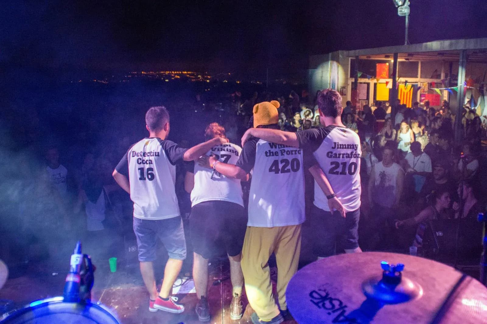 Els Kremats de espaldas en escenario nocturno con camisetas numeradas Detectiu Coca y Jimmy Garrafon ante publico de festival