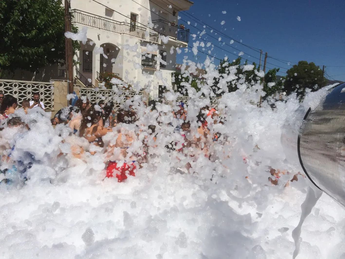 Ola de espuma blanca arrasando una piscina improvisada frente a casa rural con balcón de hierro forjado bajo cielo azul