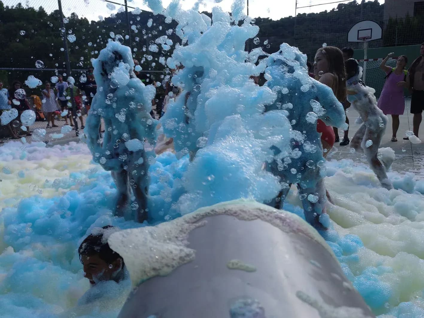 Niños jugando entre olas de espuma azul, blanca y rosa en pista deportiva con canasta de baloncesto, fiesta de la espuma