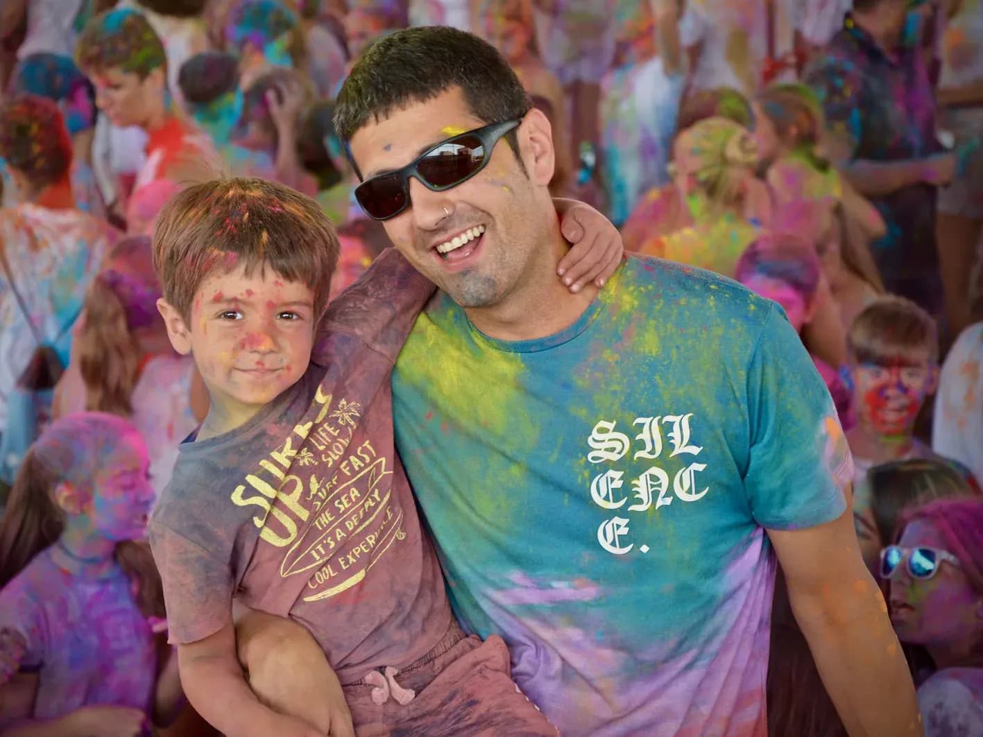 Padre con gafas de sol abrazando a un nino cubiertos de polvos de colores durante la fiesta Holi