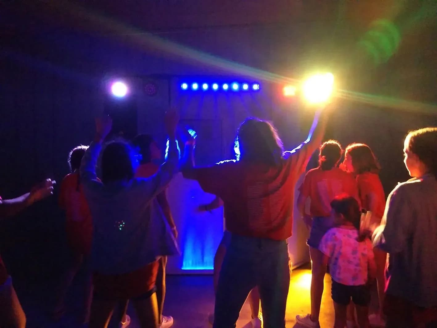 Grupo de invitados con brazos en alto bailando en sala con escenario iluminado de azul y focos de halo amarillos