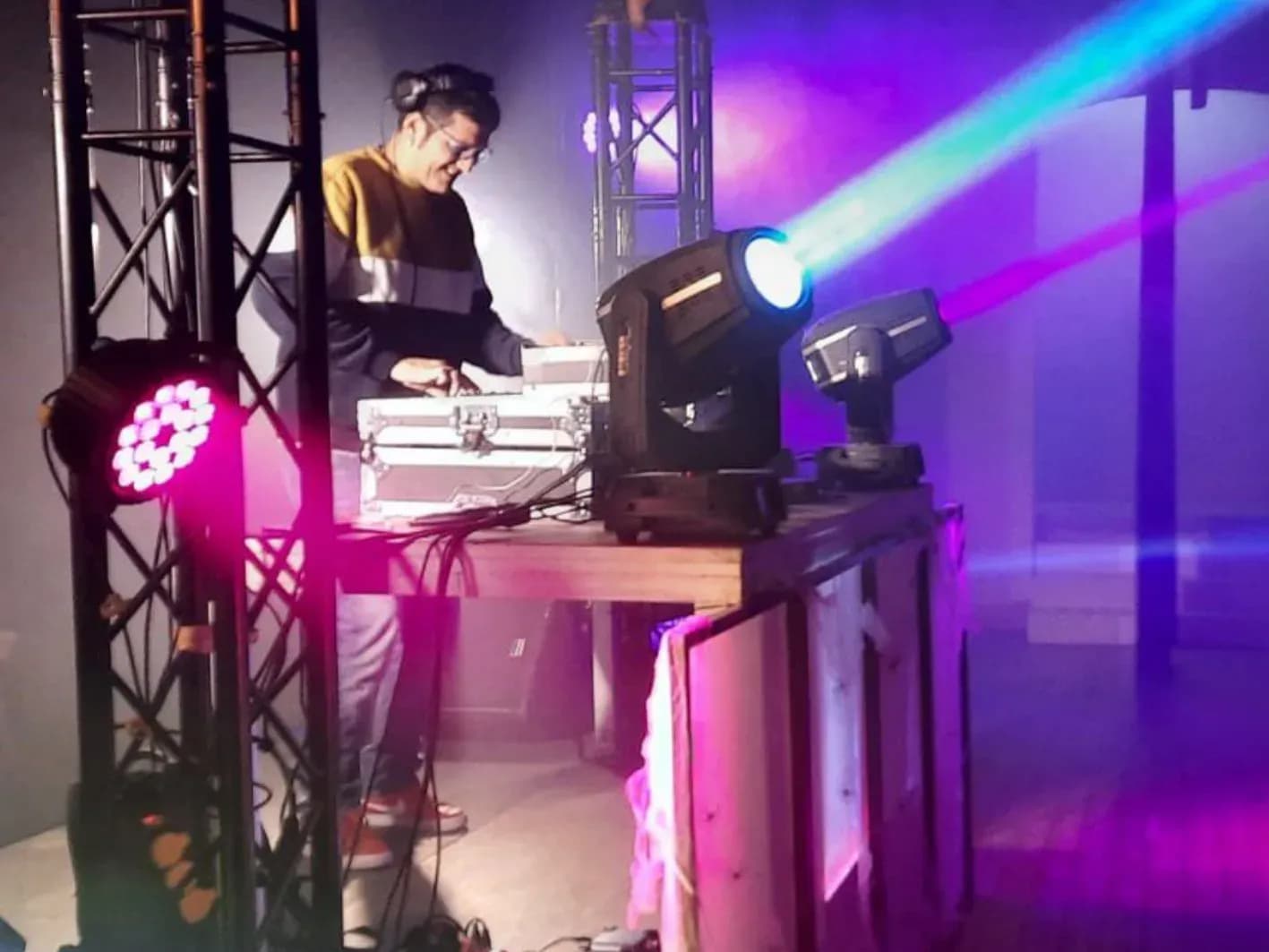 DJ con auriculares y jersey trabajando entre truss con focos LED rosa, luz láser azul y cabezas móviles en cabina