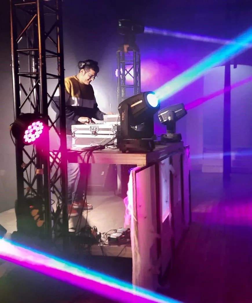 DJ con auriculares y sudadera pinchando en cabina con estructura truss, focos moviles y laseres de colores