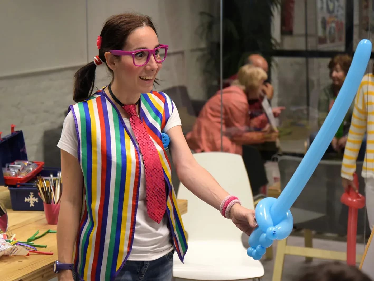 Jessica animadora con coletas, gafas magenta y chaleco de rayas modelando espada hinchable azul en taller infantil