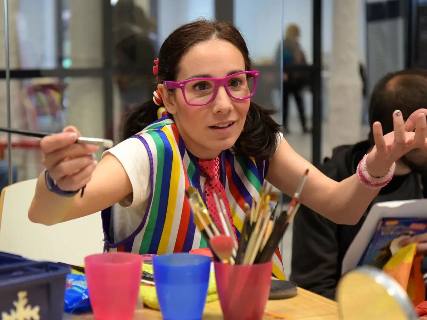 Jessica con gafas magenta y chaleco arcoíris pintando con pincel a una niña en taller de maquillaje de fantasía infantil