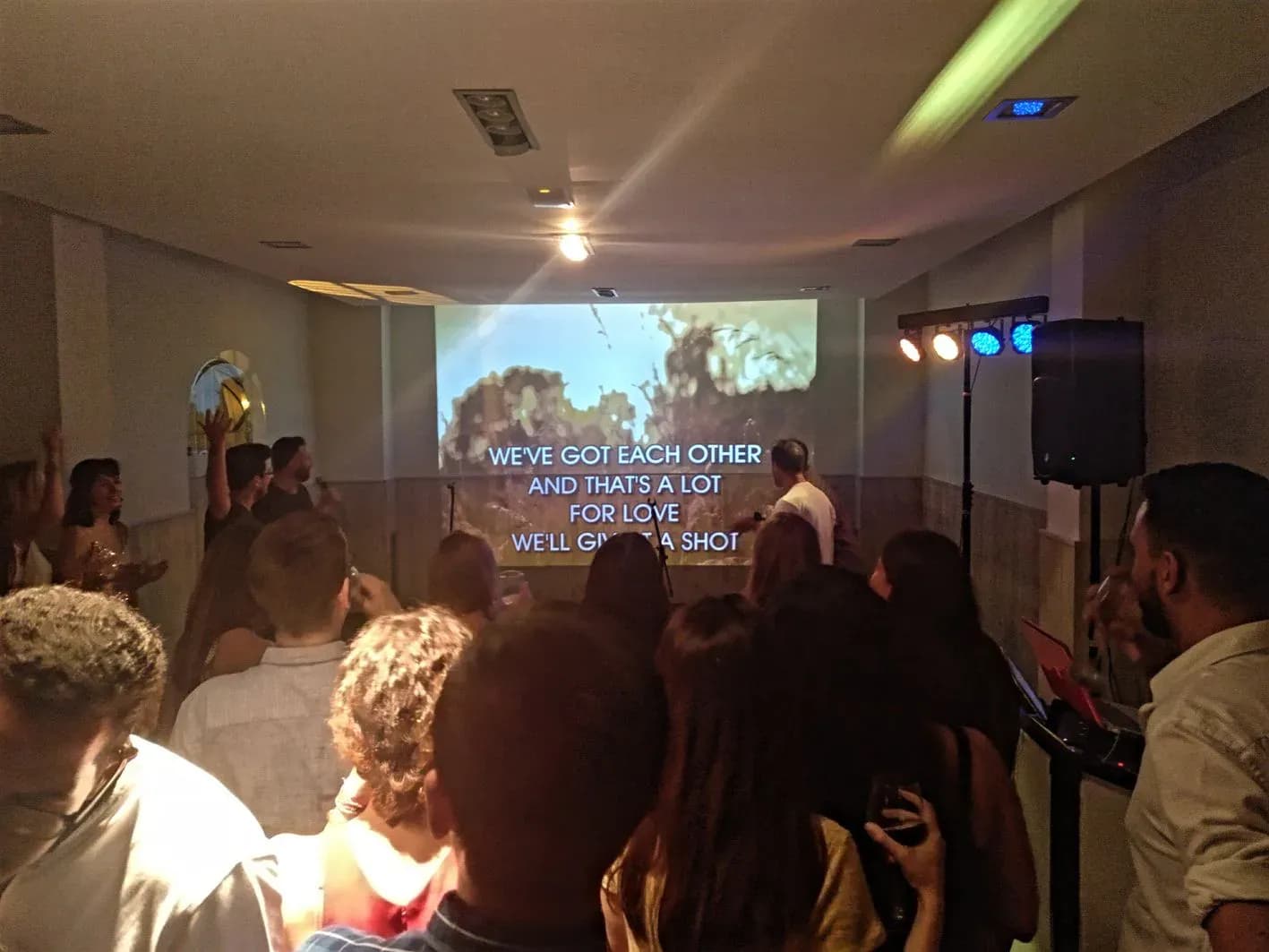 Grupo bailando frente a pantalla de karaoke con letra de canción y altavoces con focos azules en fiesta en local de eventos