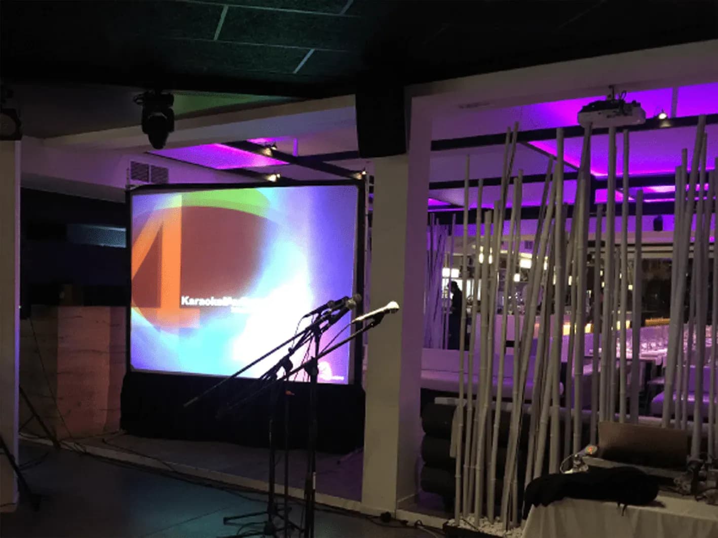 Pantalla de karaoke iluminada en violeta con micrófonos en pie y luz lateral lila junto a sala con mesas vestidas