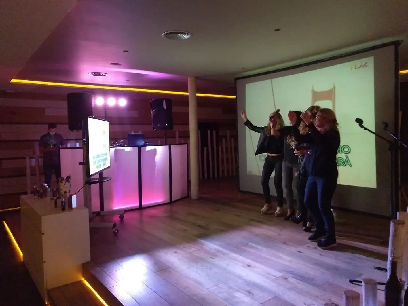 Grupo cantando en karaoke ante pantalla con altavoces y cabina de DJ retroiluminada de rosa fucsia en sala con suelo de madera