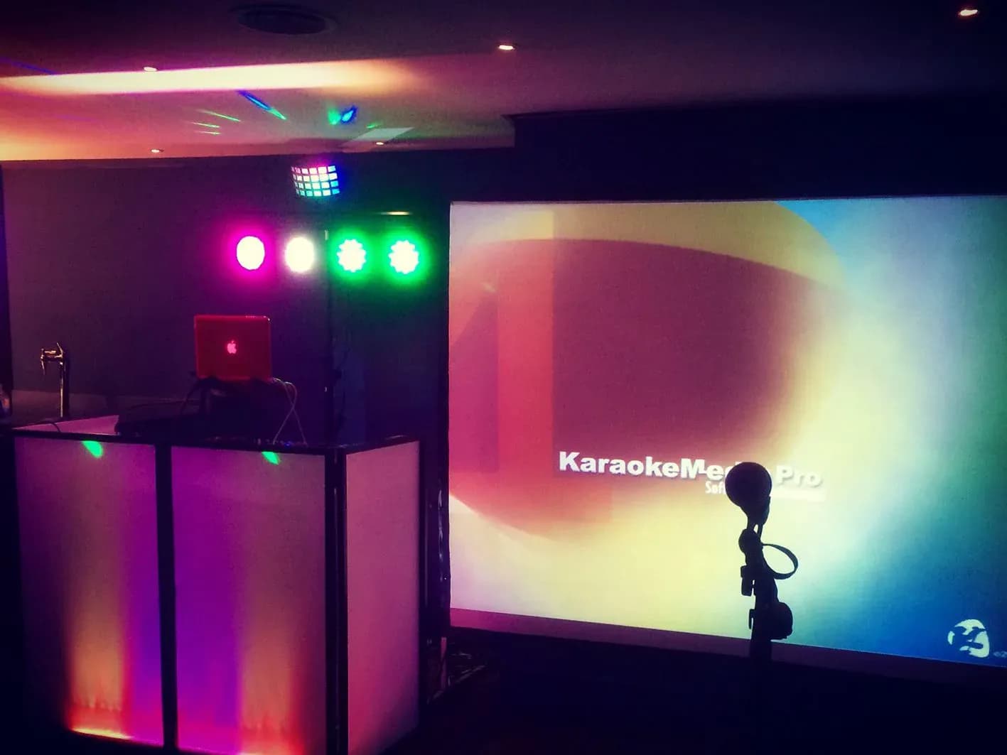 Cabina de karaoke iluminada con focos de colores y pantalla proyectando KaraokeMedia Pro junto al microfono