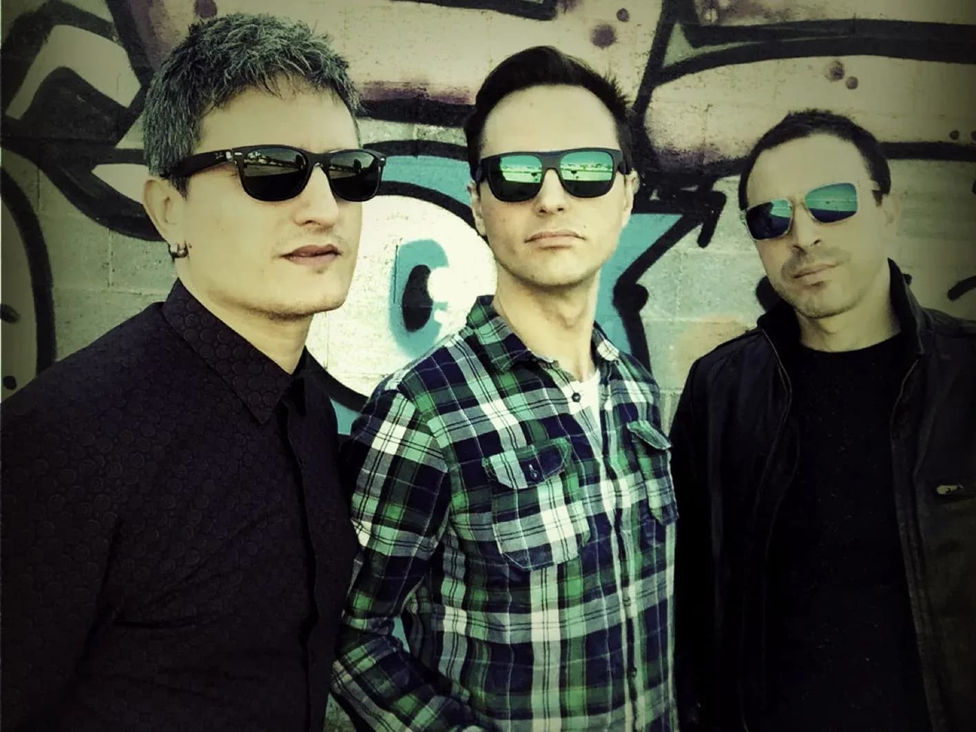 Tres integrantes de Mind Trick con gafas de sol y camisa de cuadros ante pared con grafitis, retrato urbano de la banda