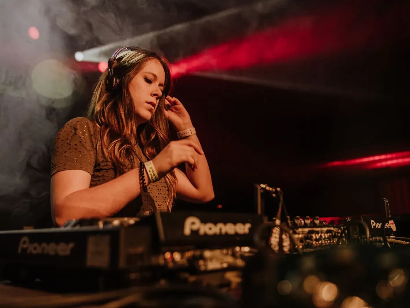 Mireia DG con auriculares y top de lunares ajustándose la pulsera tras mesa Pioneer entre humo y luces rojas