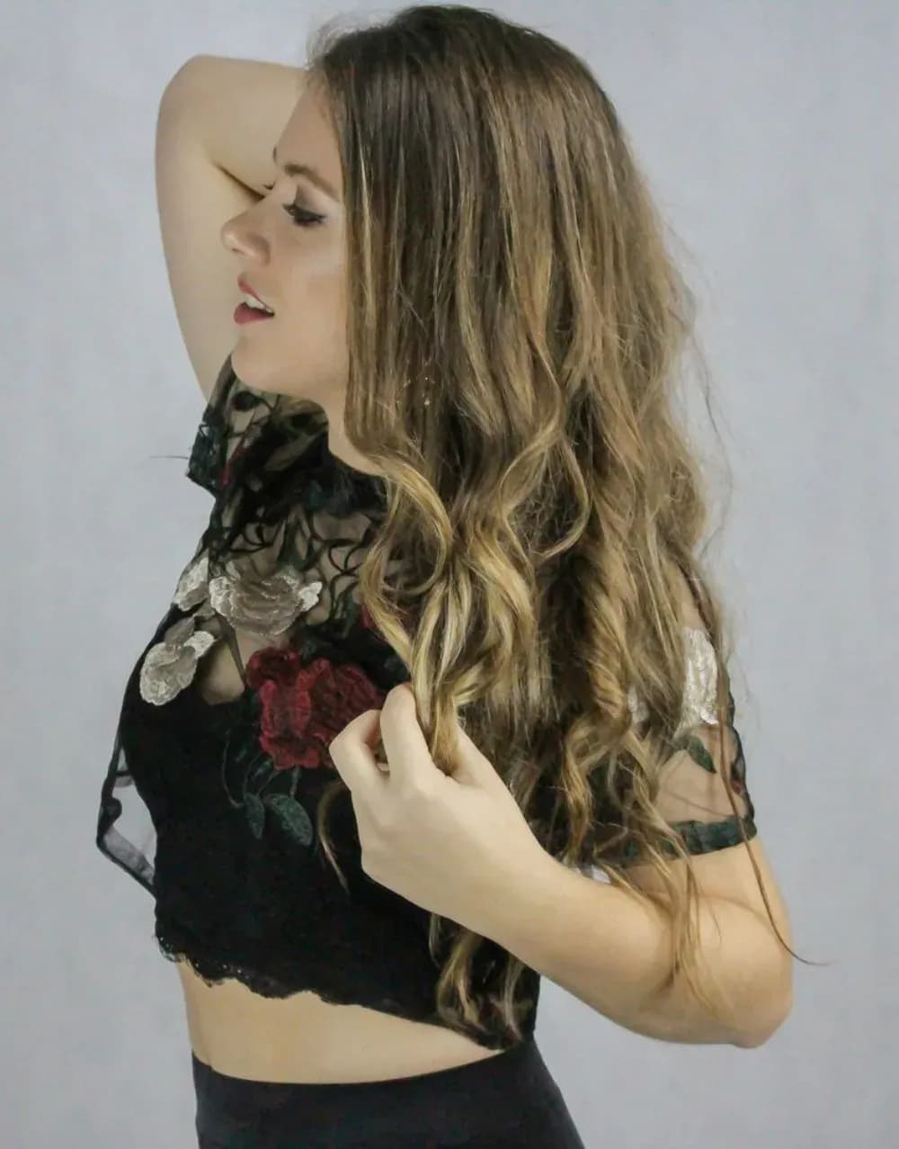 Retrato lateral de Mireia DG con top de encaje floral negro y melena rubia ondulada, sesión promocional sobre fondo gris