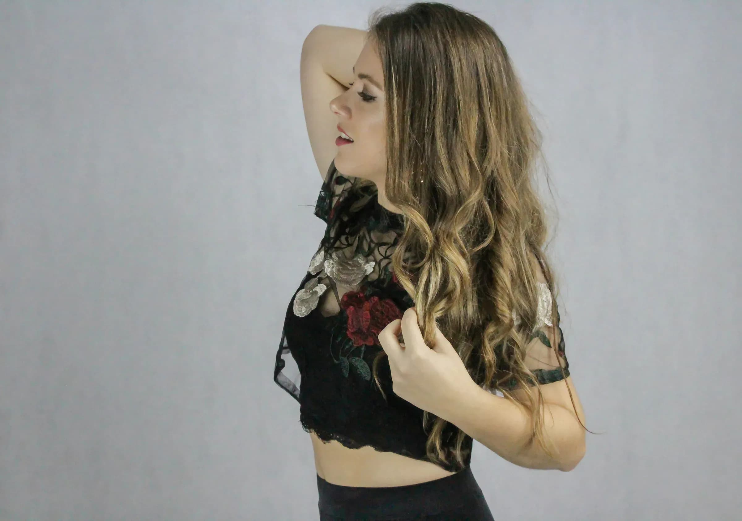 Mireia DG de perfil con melena ondulada y top de encaje negro con flores bordadas sobre fondo gris claro