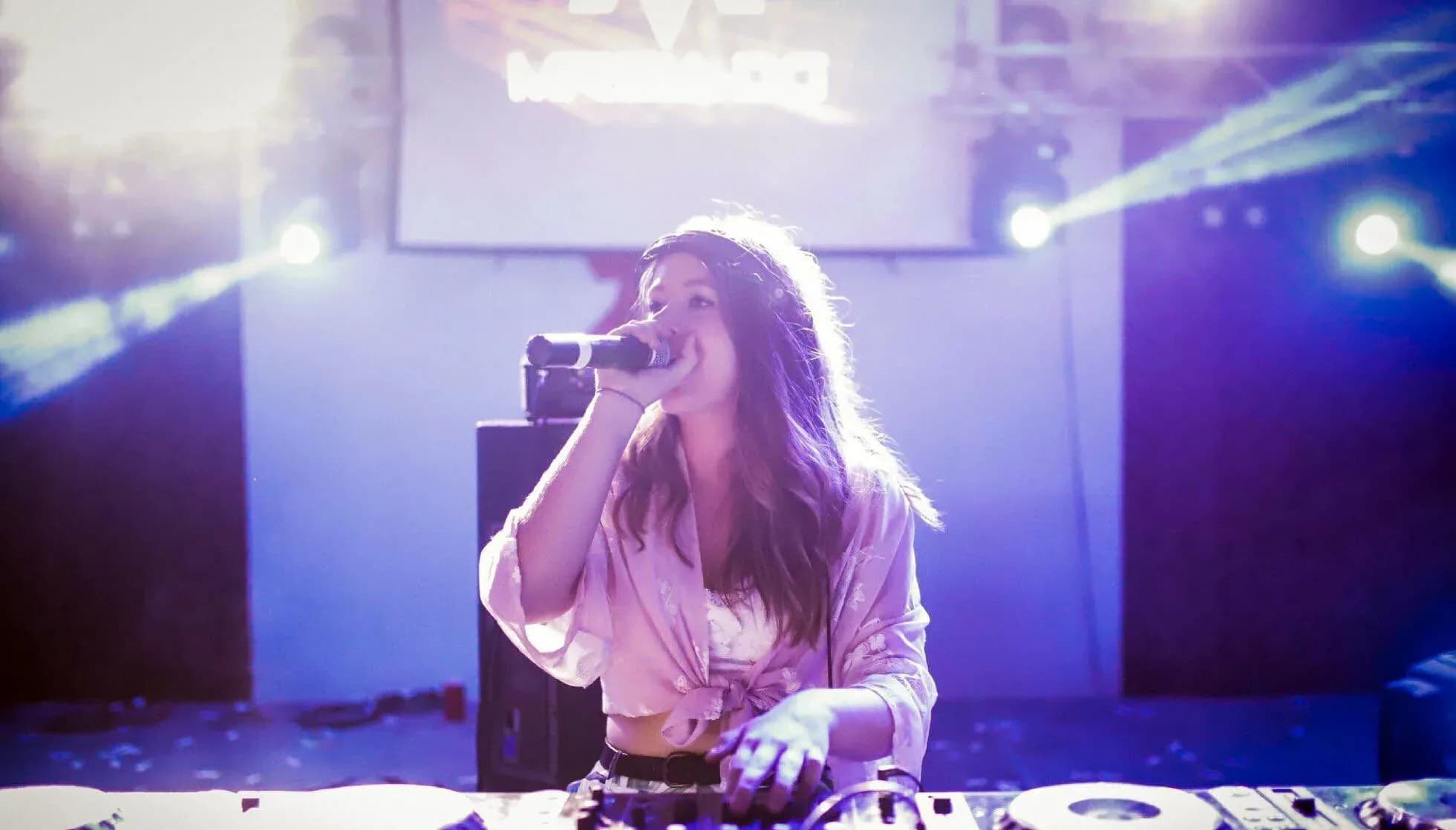 DJ Mireia rubia con auriculares, cazadora de cuero negra y top blanco posando sobre fondo claro de estudio