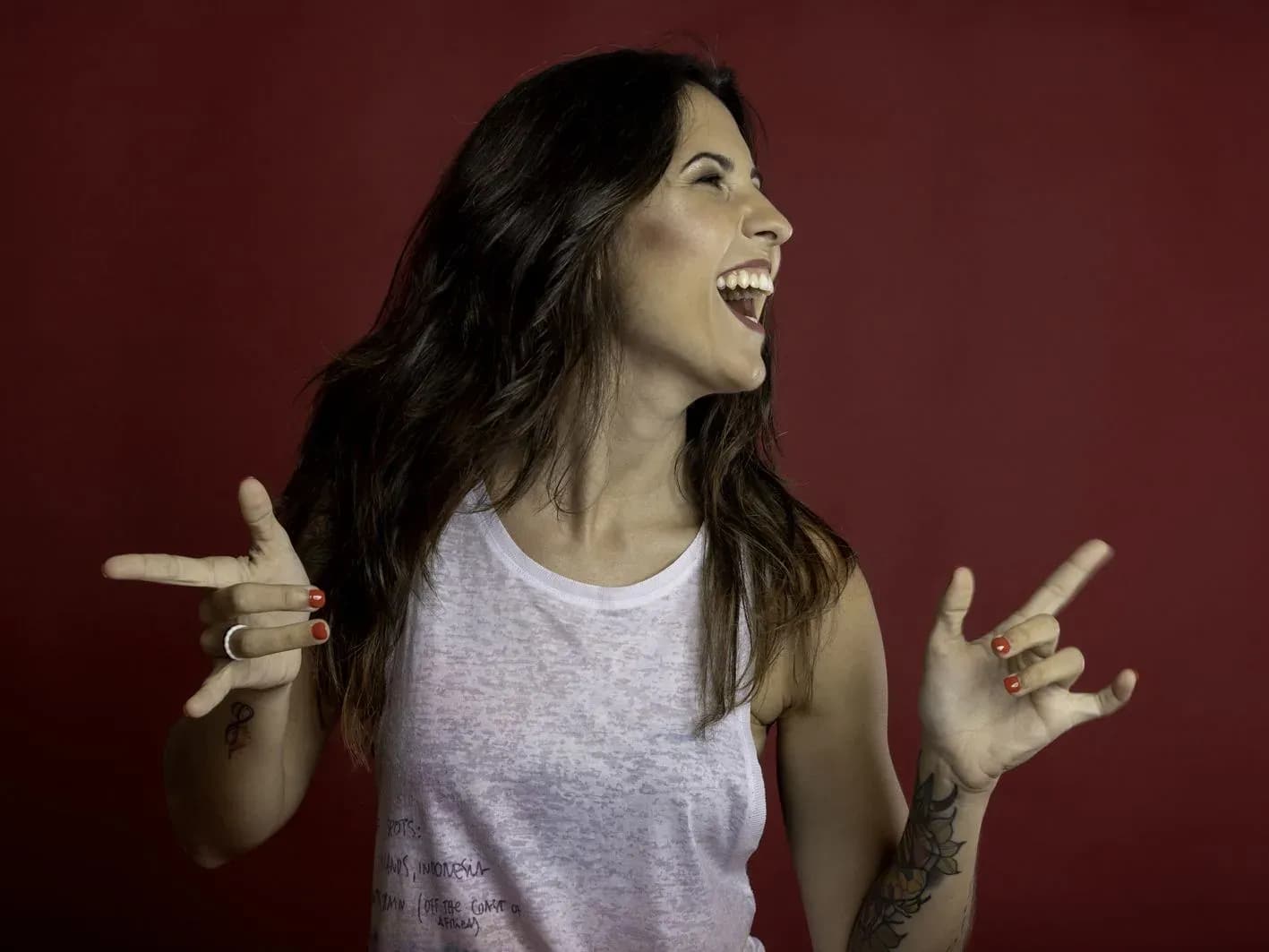 Nuria Scarp con camiseta blanca riendo y haciendo cuernos de rock con las manos sobre fondo rojo, retrato promocional