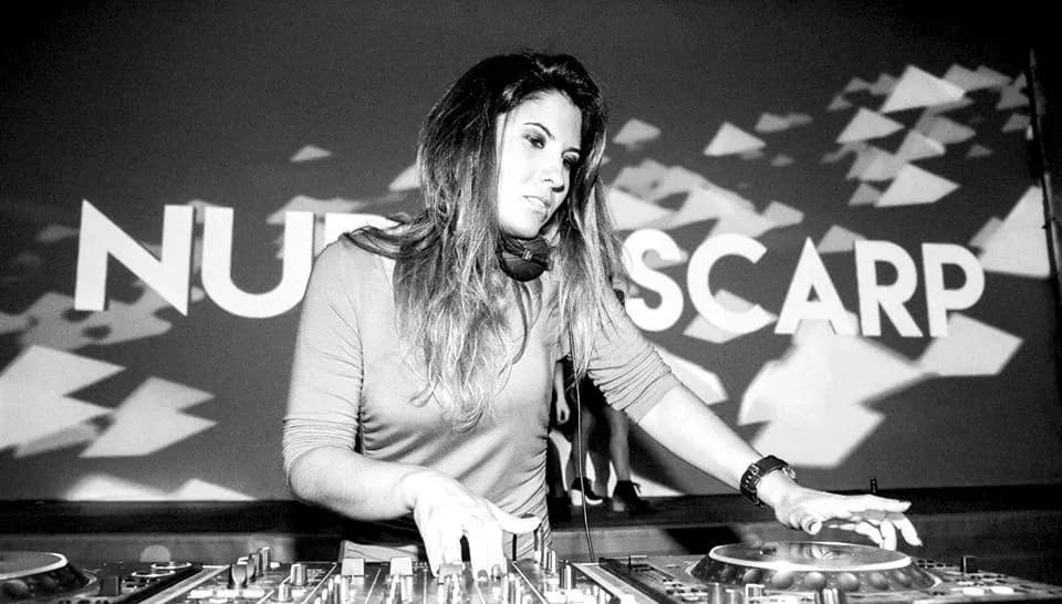 DJ Nuria Scarp pinchando con auriculares al cuello en cabina, fotografia en blanco y negro con logo de fondo
