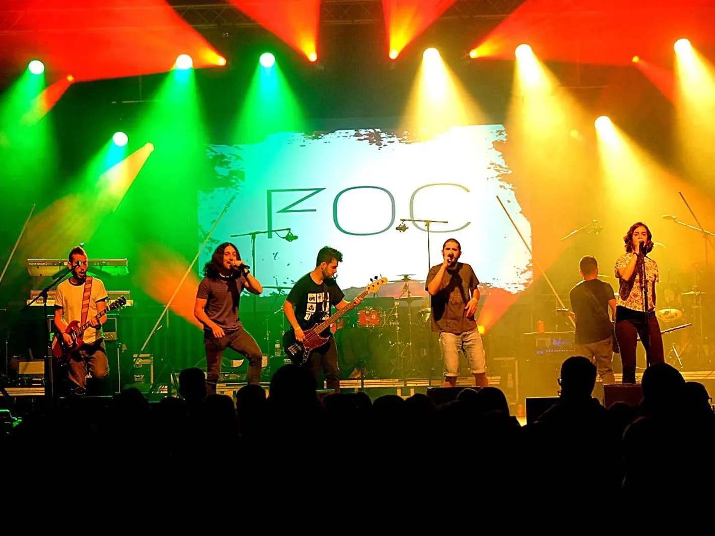 Banda Foc en directo con cinco músicos sobre escenario iluminado de verde, rojo y amarillo ante proyección con su logotipo