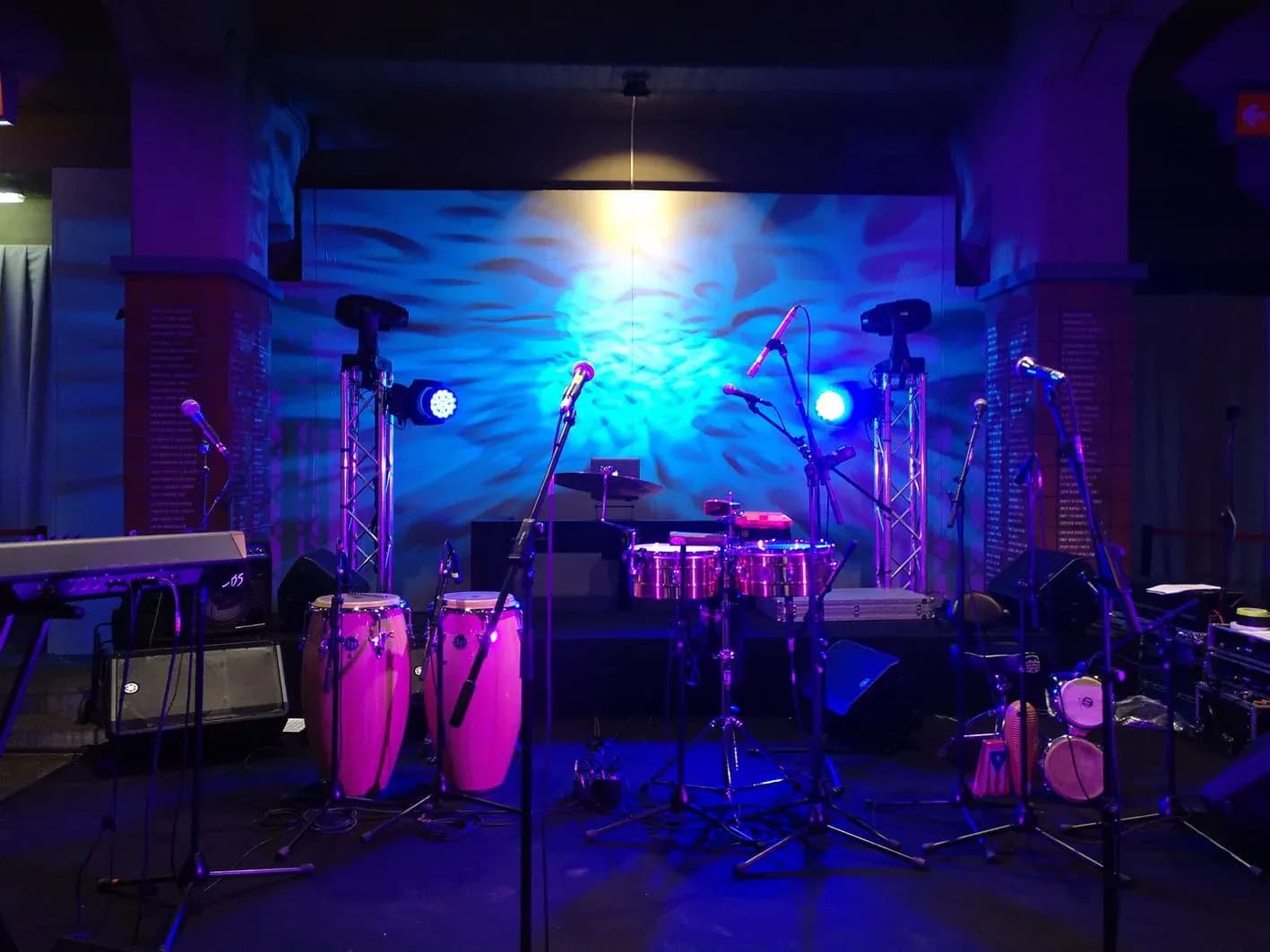 Escenario montado con bateria, congas rosas y trusses iluminados con focos azules y violetas para concierto en sala