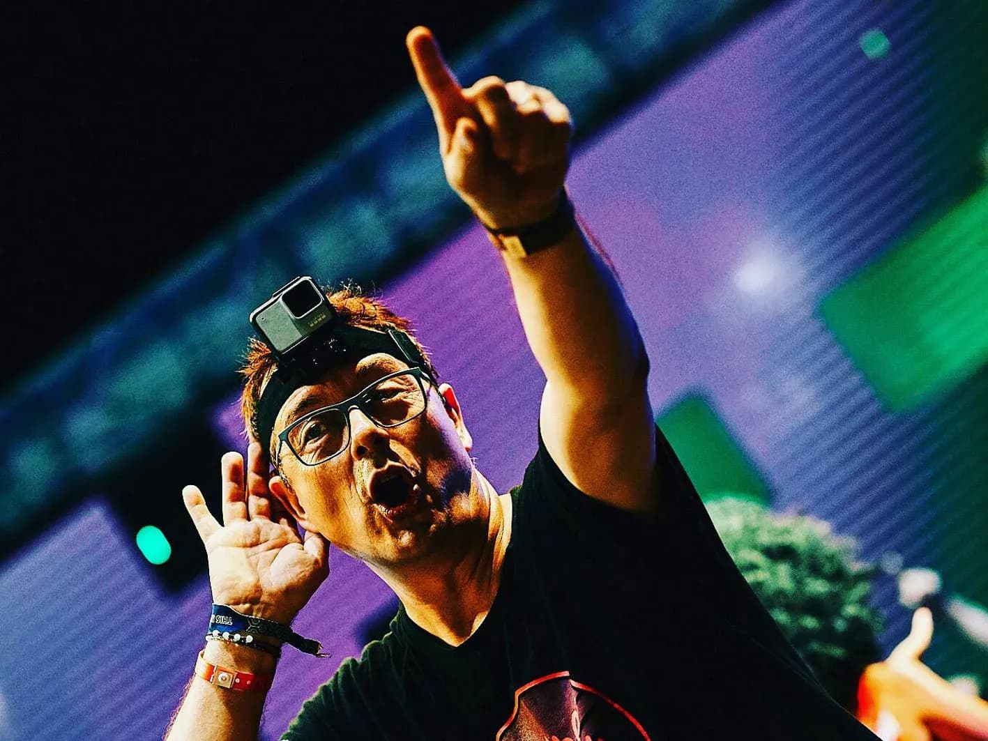 Toni Peret con GoPro en la cabeza señalando al cielo y aguantando el auricular en concierto al aire libre con pantalla LED