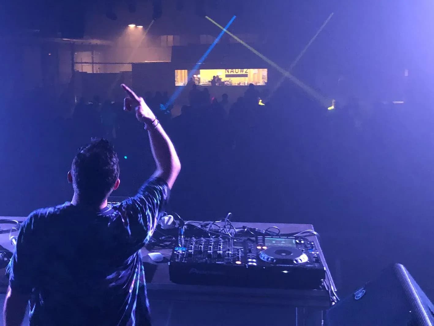 Toni Peret de espaldas señalando al público desde la cabina del festival NAU#2 con láseres azules y verdes en sala