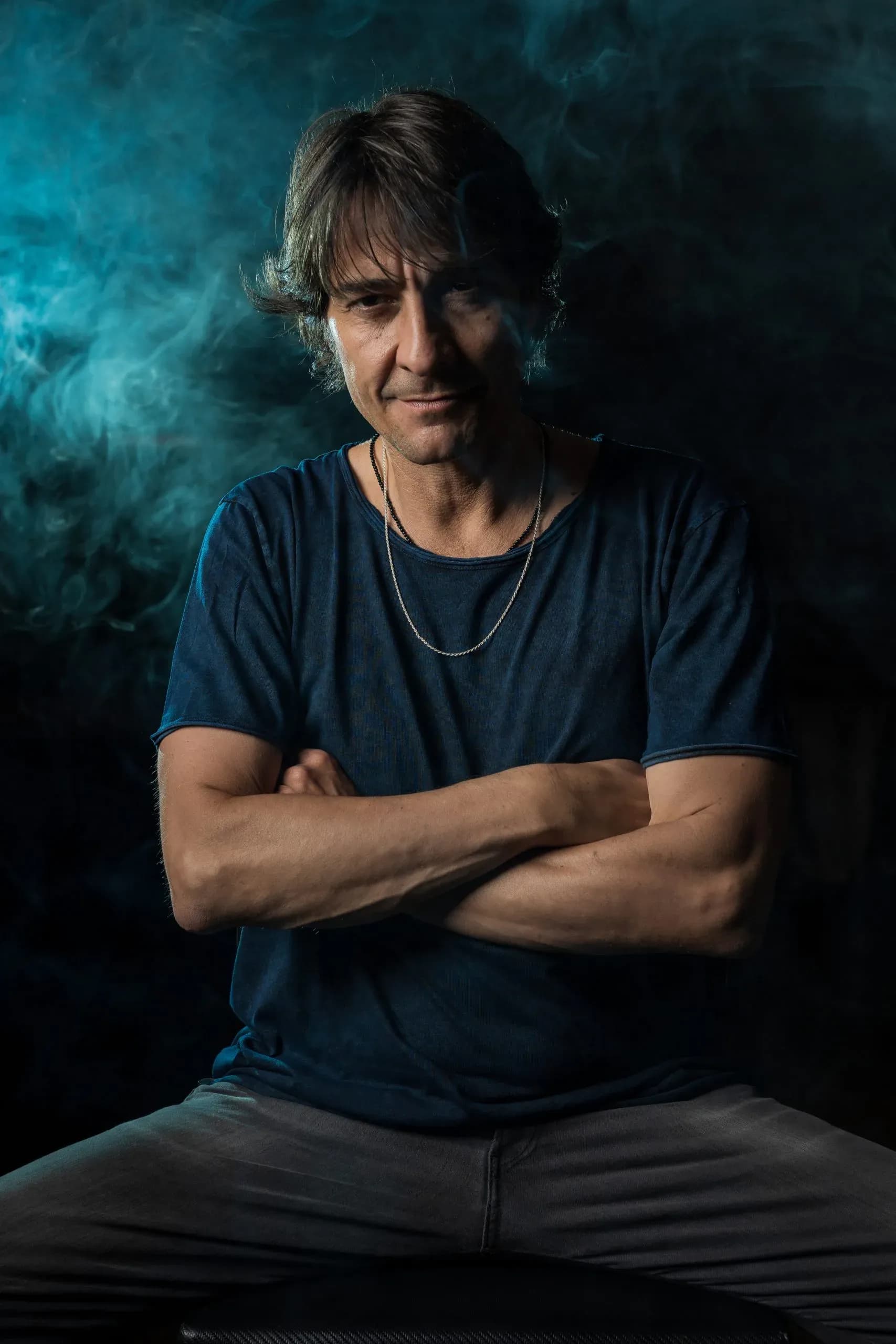 Abel Almena sentado con los brazos cruzados sobre fondo oscuro y humo turquesa, retrato promocional del DJ residente