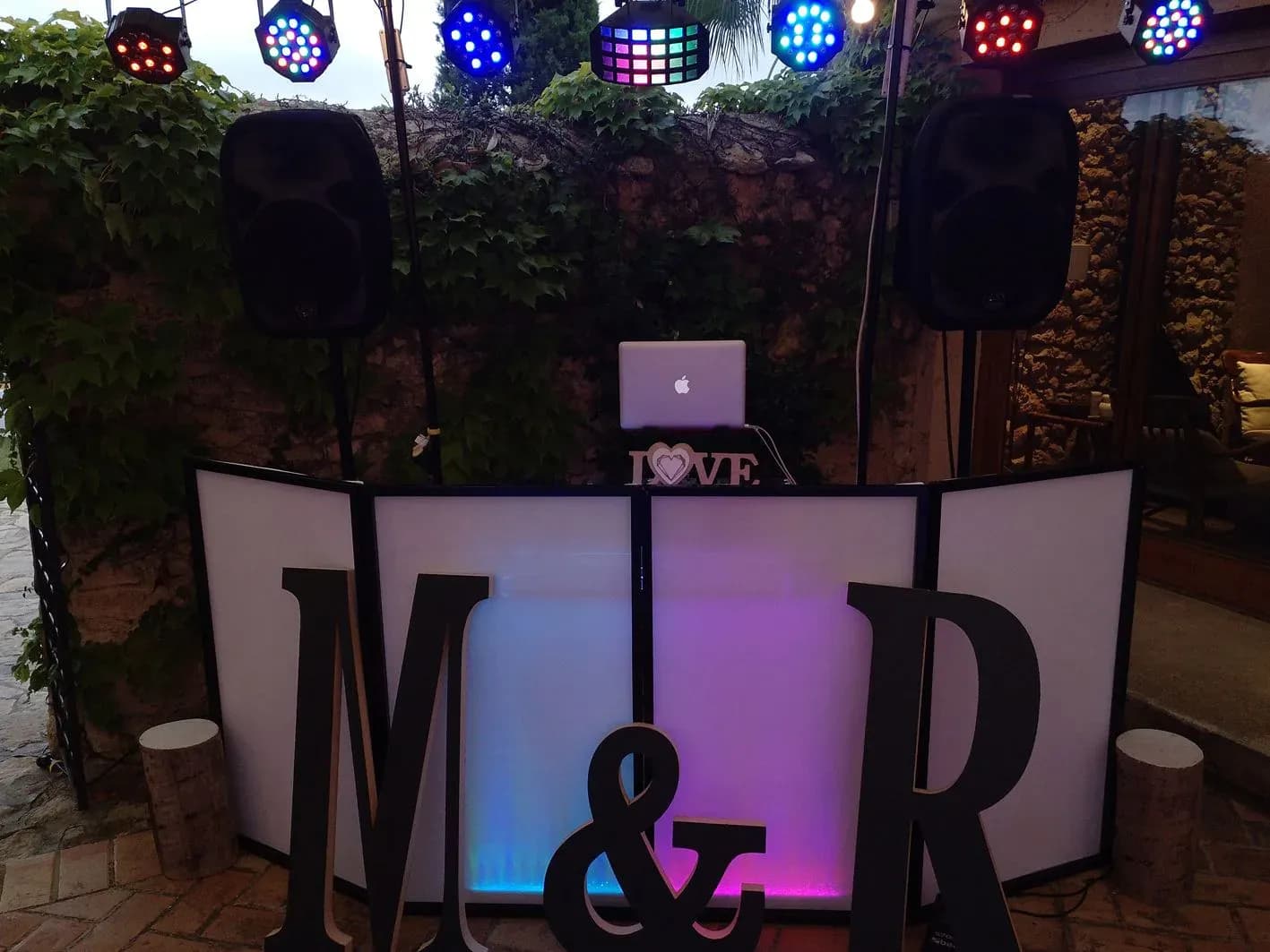 Cabina de DJ para boda con letras gigantes M&R, altavoces, focos LED y Macbook con cartel LOVE sobre muro de hiedra