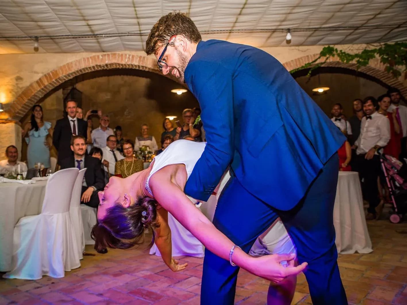 Novio inclinando a la novia en figura de baile durante el baile nupcial bajo arcos de ladrillo con invitados aplaudiendo