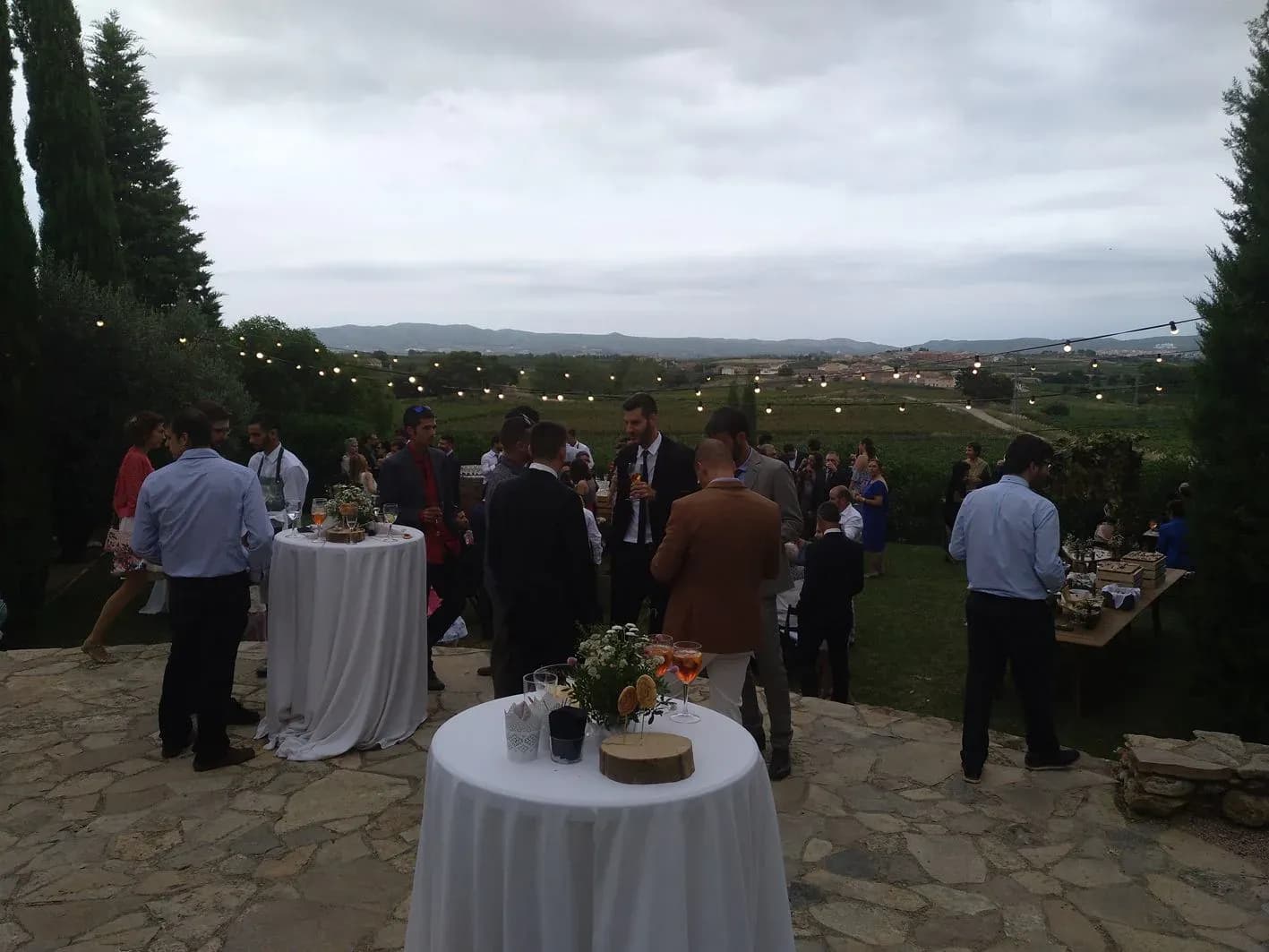 Aperitivo de boda al aire libre con mesas vestidas, guirnaldas de luces, invitados charlando y vistas a viñedos