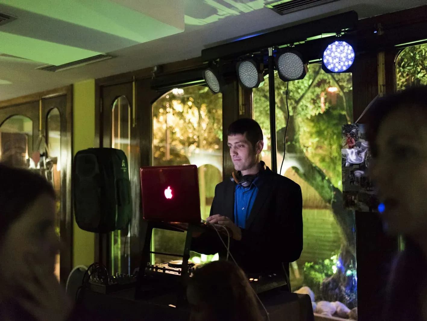DJ en boda pinchando con portatil rojo y americana oscura en sala acristalada con focos blancos colgantes
