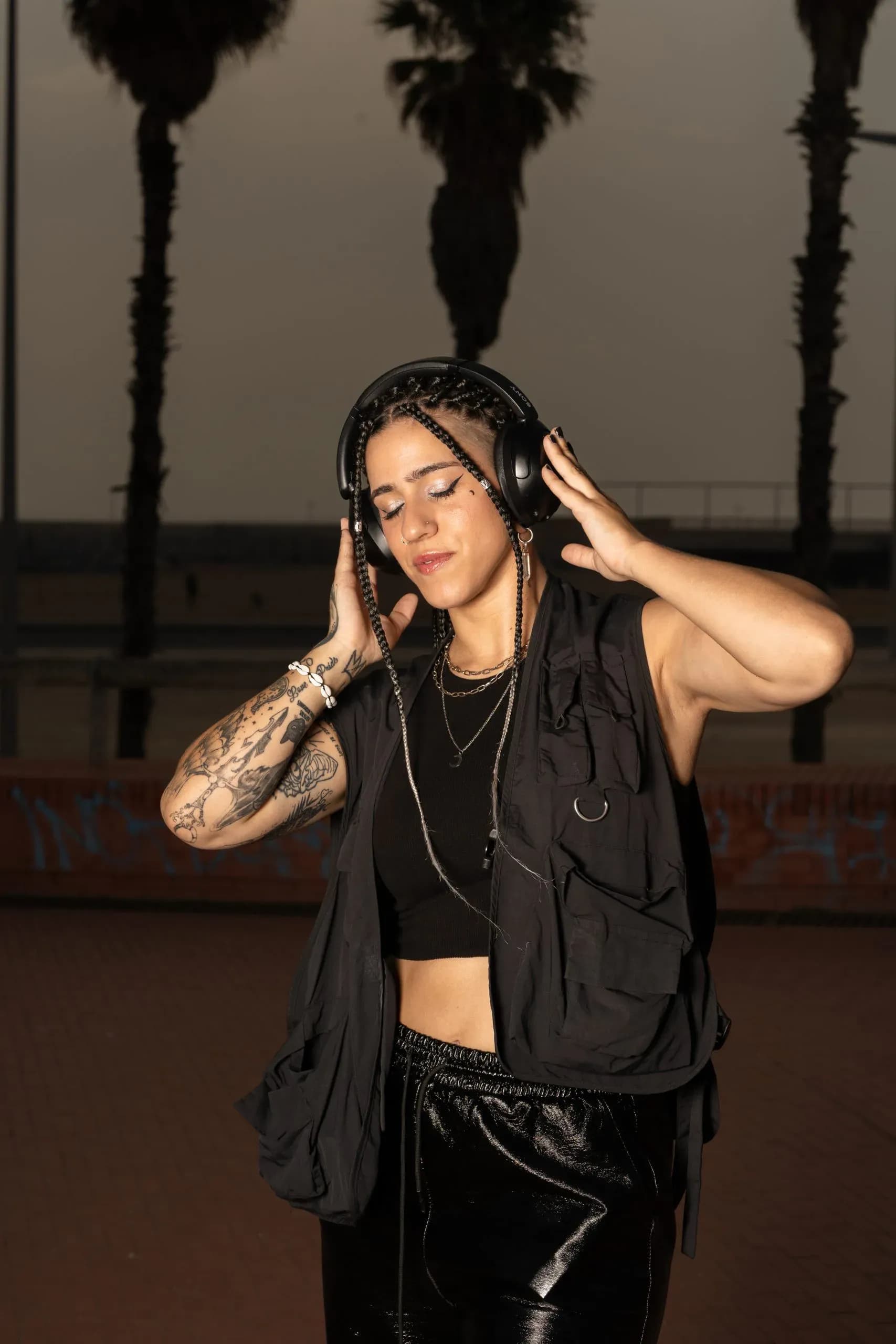 Carla Nisha con auriculares y chaleco negro frente a palmeras al atardecer, DJ urbana barcelonesa de hip-hop y dembow