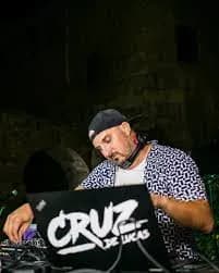 DJ Cruz de Luca con gorra negra y camisa estampada pinchando tras cartel propio en sesión nocturna al aire libre