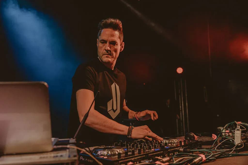 DJ Iván Jiménez con camiseta del logo IJ mezclando con laptop y mesa Pioneer bajo humo azul en escenario de festival