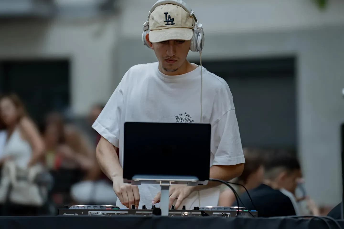 DJ Kolom con gorra LA blanca y camiseta clara pinchando con MacBook y mesa Pioneer DJ en evento al aire libre