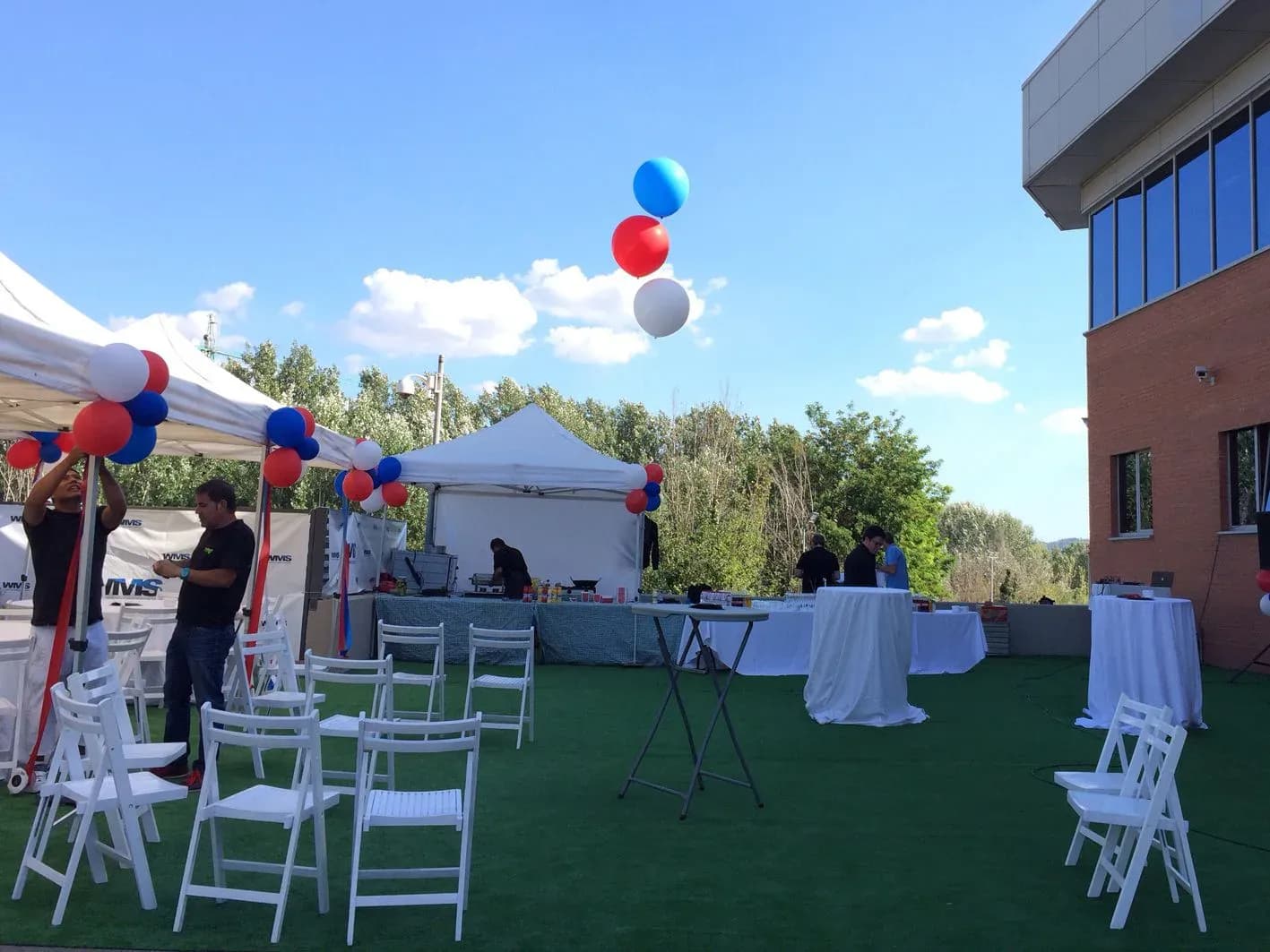 Family day de empresa con globos rojo, blanco y azul, sillas plegables y carpa de catering sobre césped al sol