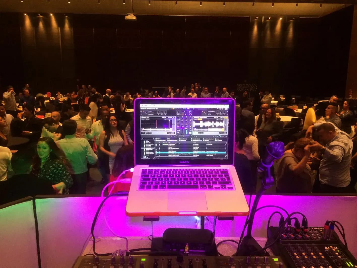Cabina de DJ con portatil iluminado en violeta y publico bailando al fondo en fiesta de empresa