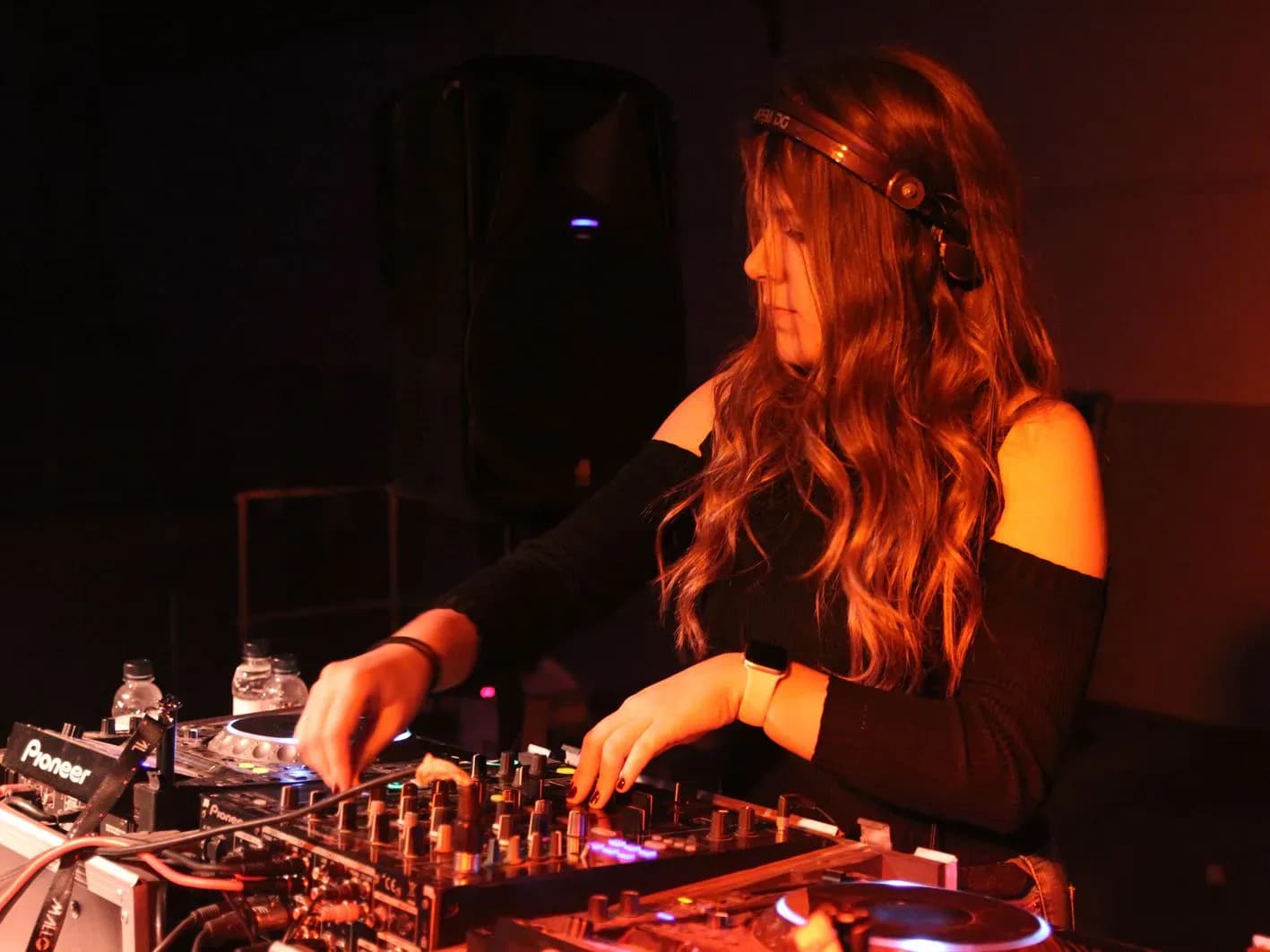 Mireia DG con auriculares Pioneer y top negro manipulando mesa Pioneer bajo luz cálida en cabina de festival